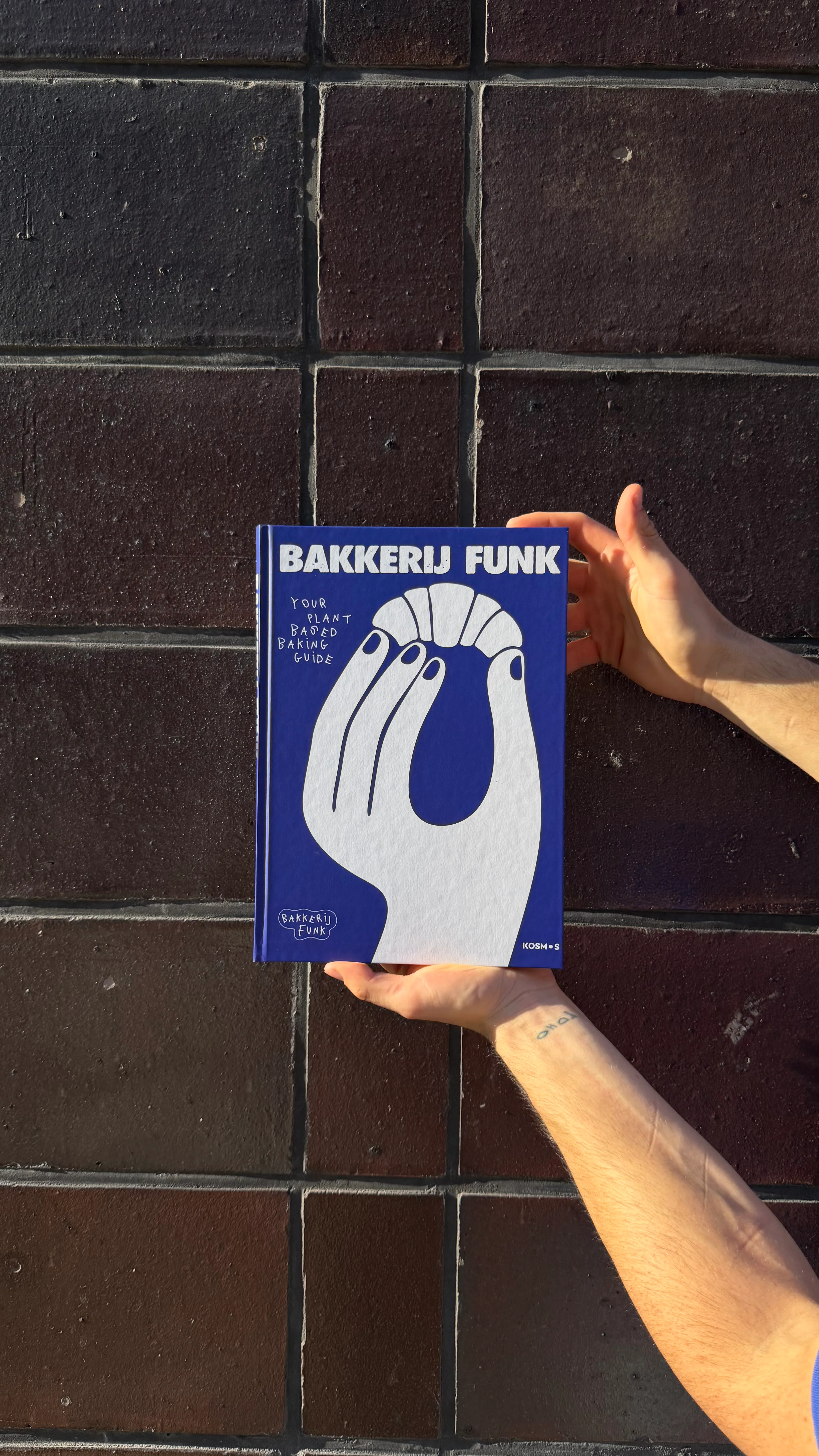 Hands holding the Bakkerij FUNK book 'Your Plant Based Baking Guide' against a dark brick wall