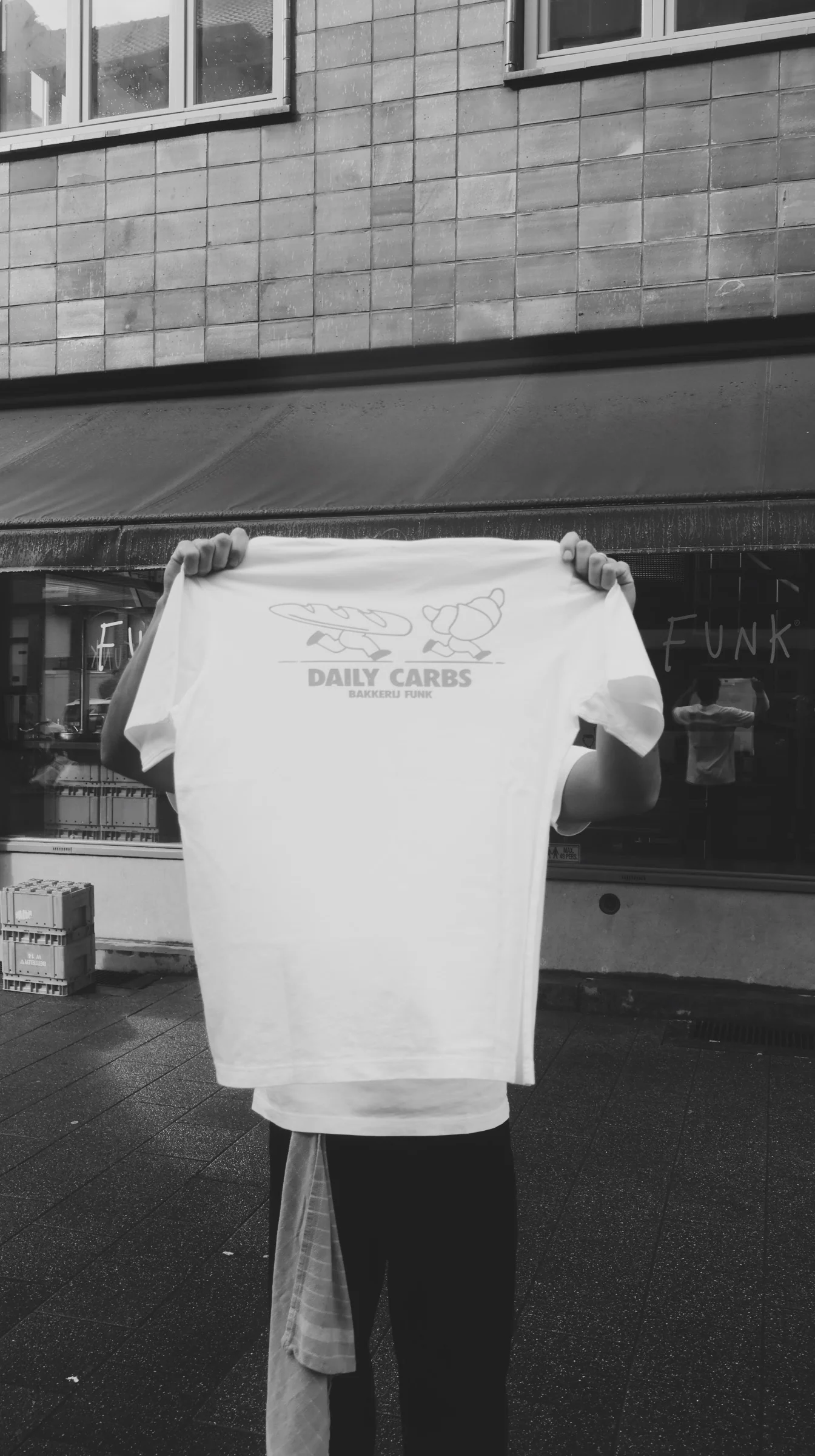 A person holding up a Bakkerij FUNK 'Daily Carbs' t-shirt in front of the FUNK shop